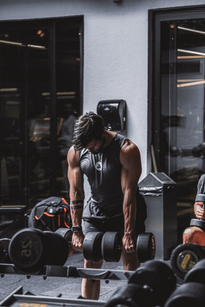 A muscular man in a tank top lifts heavy dumbbells in a gym setting.